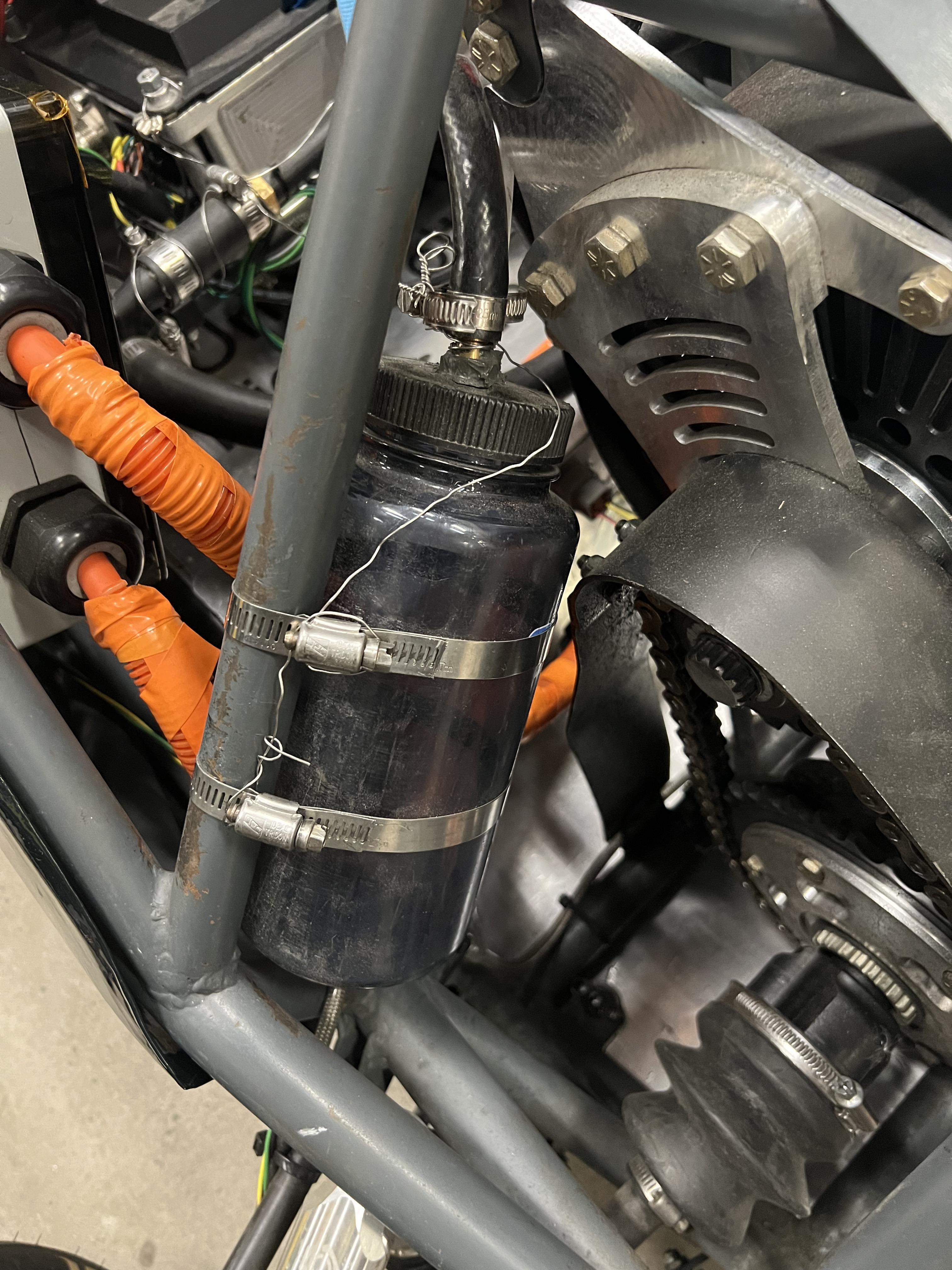 A dark plastic catch can clamped to a chassis tube with two hose clamps, with an overflow line running into its cap