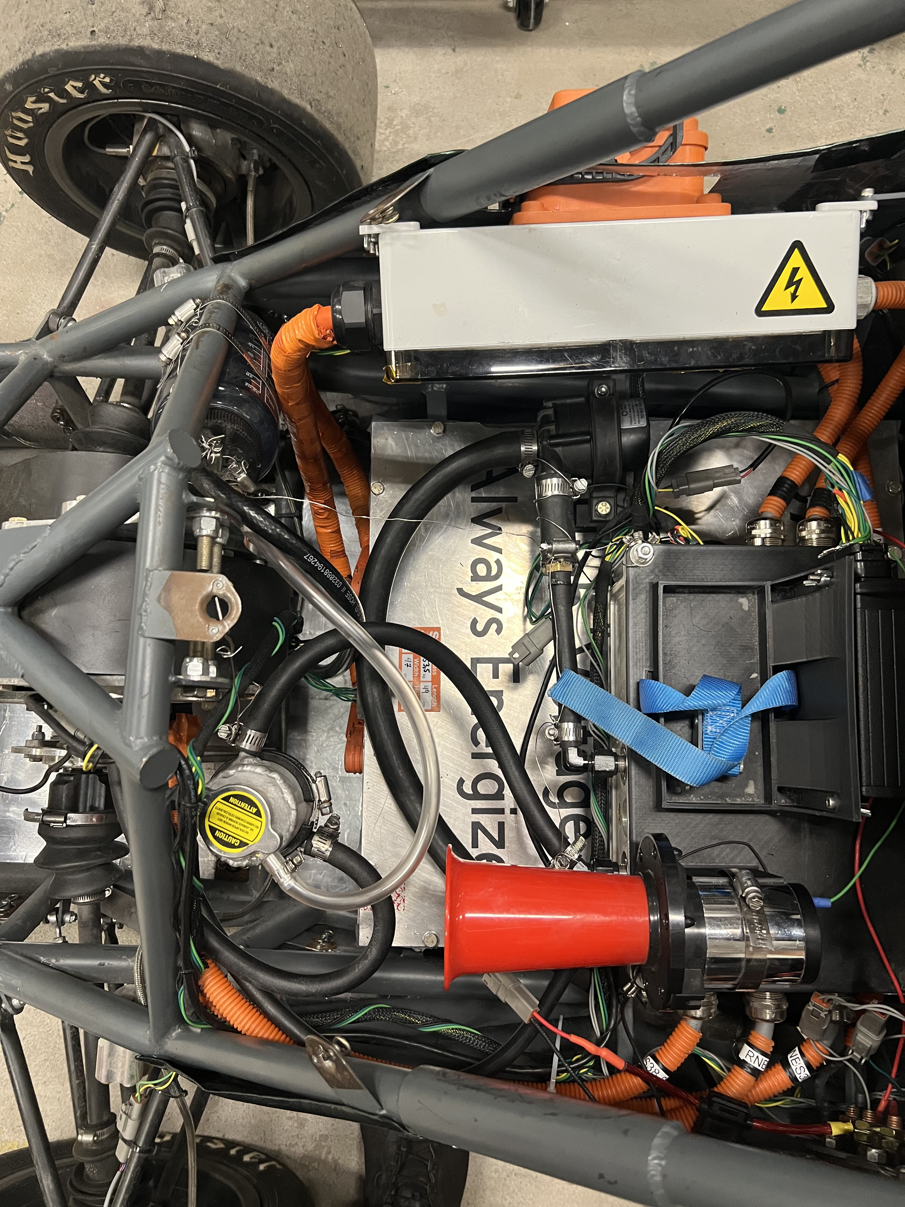 Overhead view of the rear of the car showing black coolant hoses routed between the swirl pot, the pump, and the powertrain hardware, alongside orange high-voltage cabling
