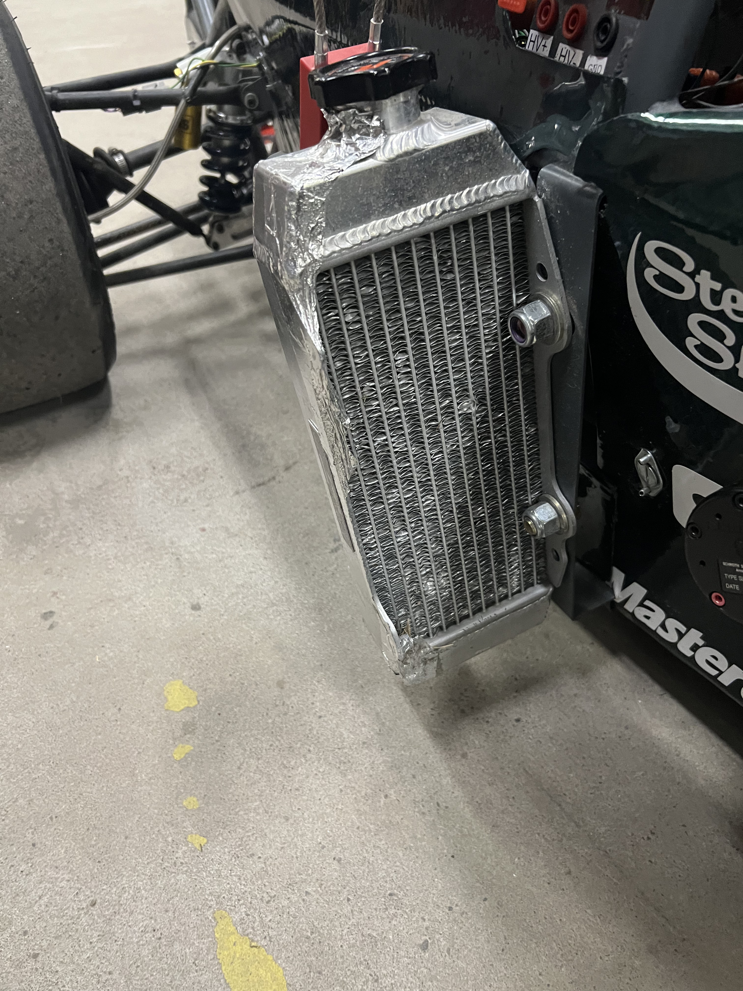 An aluminum radiator with welded end tanks and a pressure cap, bolted outboard at the side of the car