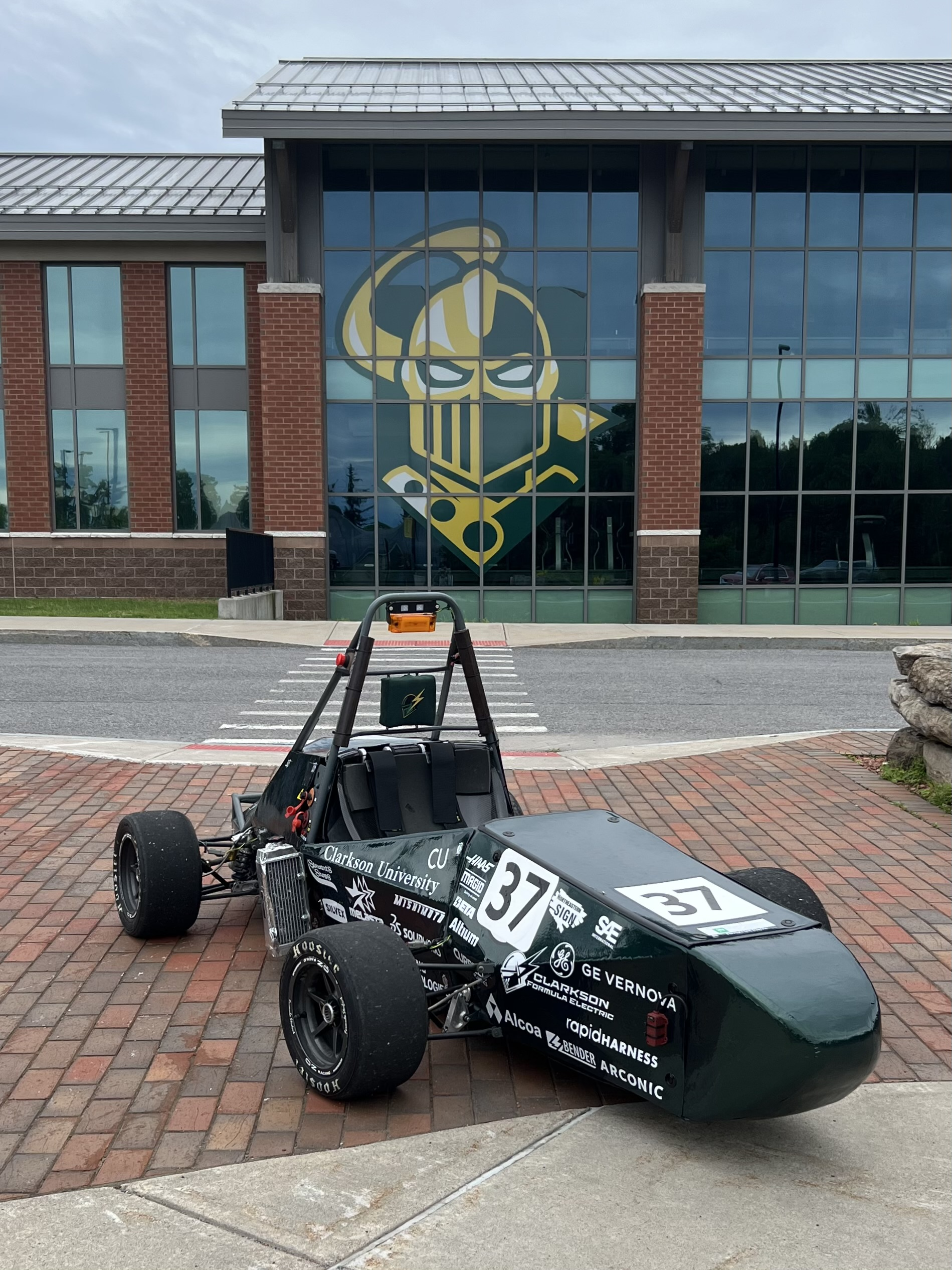 The team's dark green formula car, number 37, parked on a brick plaza outside a Clarkson University building with the Golden Knight logo in the windows behind it