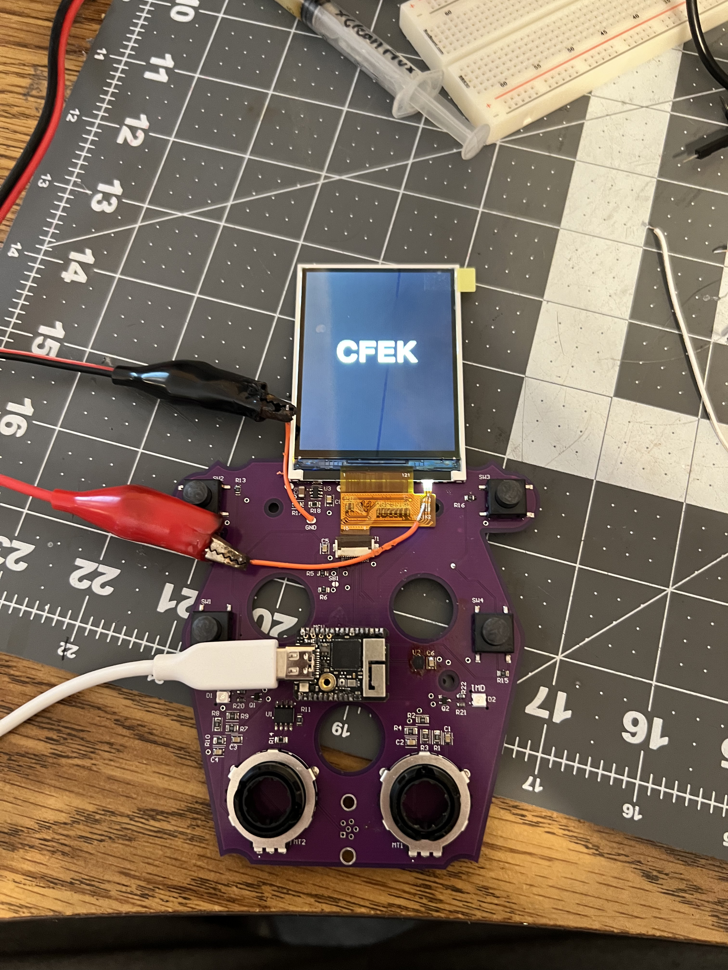 The bare dash PCB on a cutting mat during development, wired to a TFT display showing the CFEK logo, with alligator clips and a USB cable attached